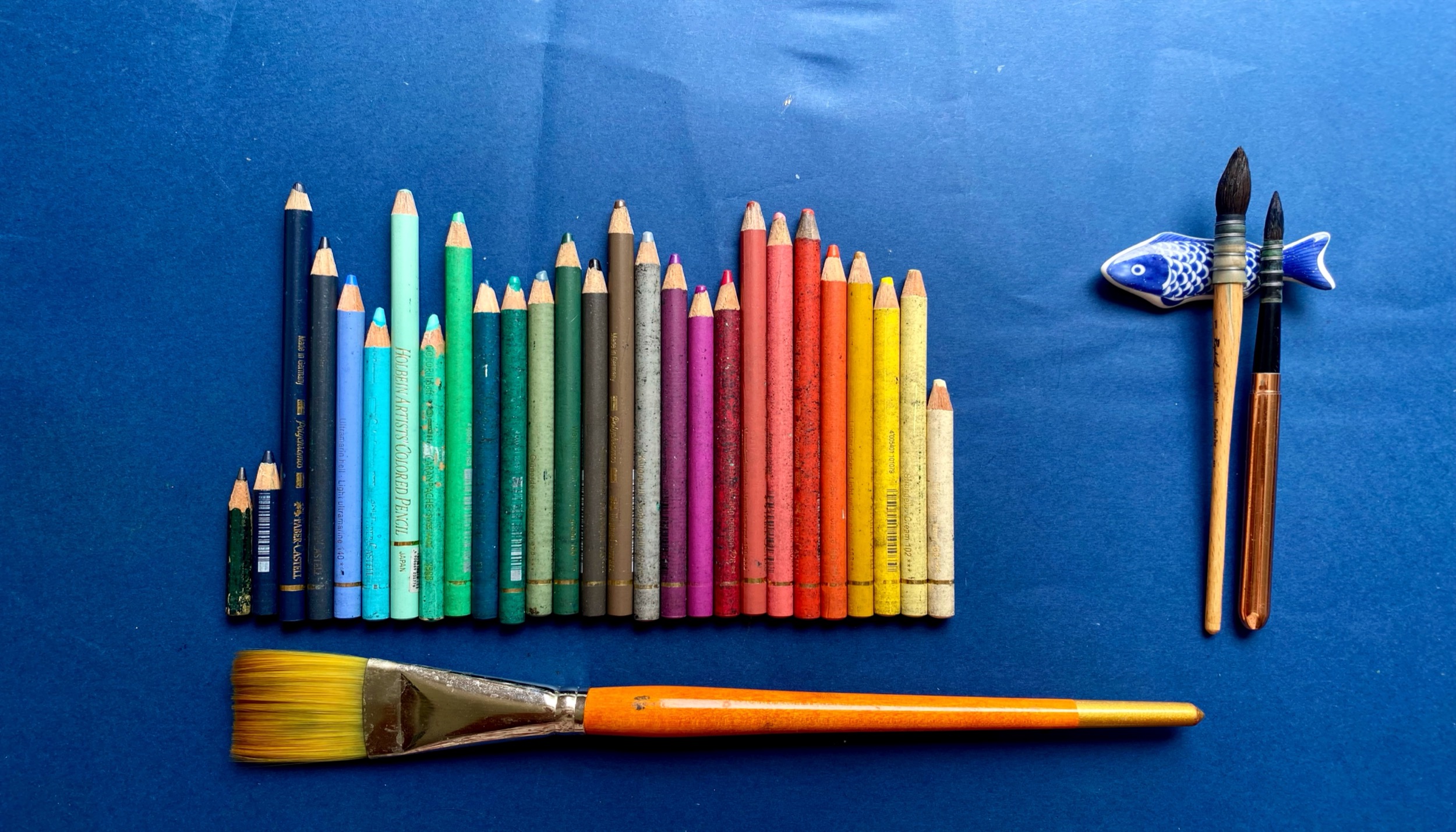 Colored pencils, brushes, and a blue ceramic fish arranged on blue paper.