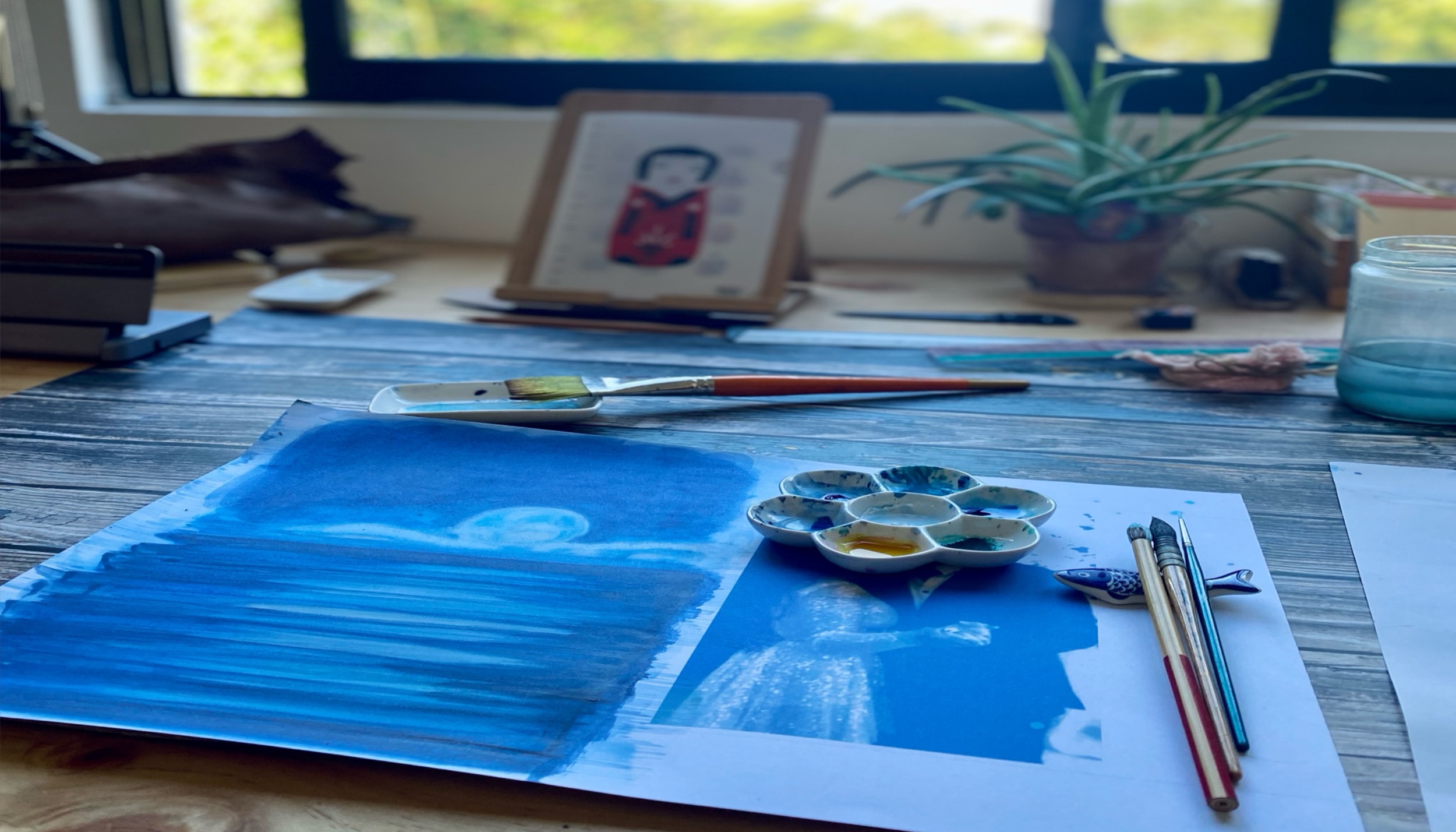 Blue watercolor painting, brushes, and studio materials on a work table.